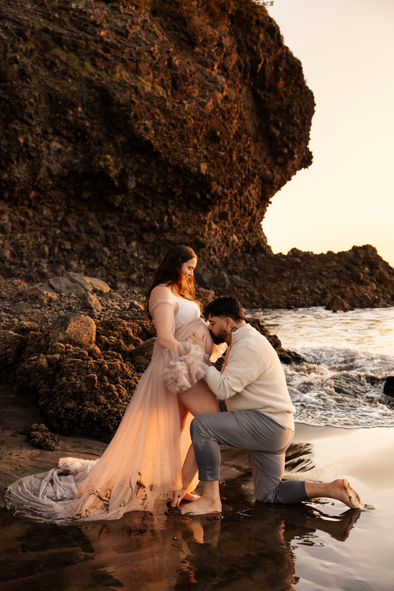 pregnant couple standing at front of the cliff at beach and dad kissing belly