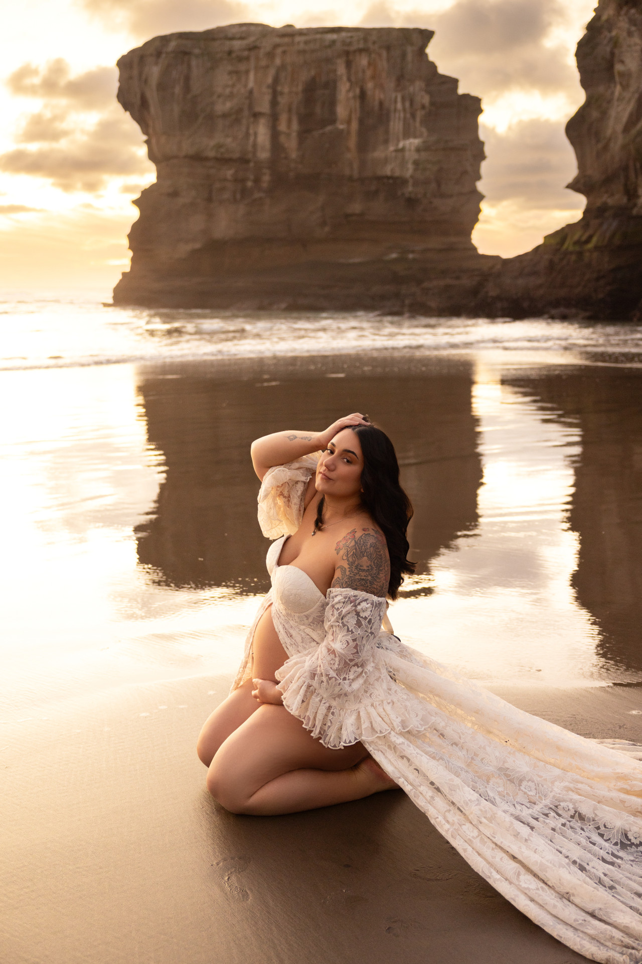 pregnant woman kneeling at muriwai beach looking in camera