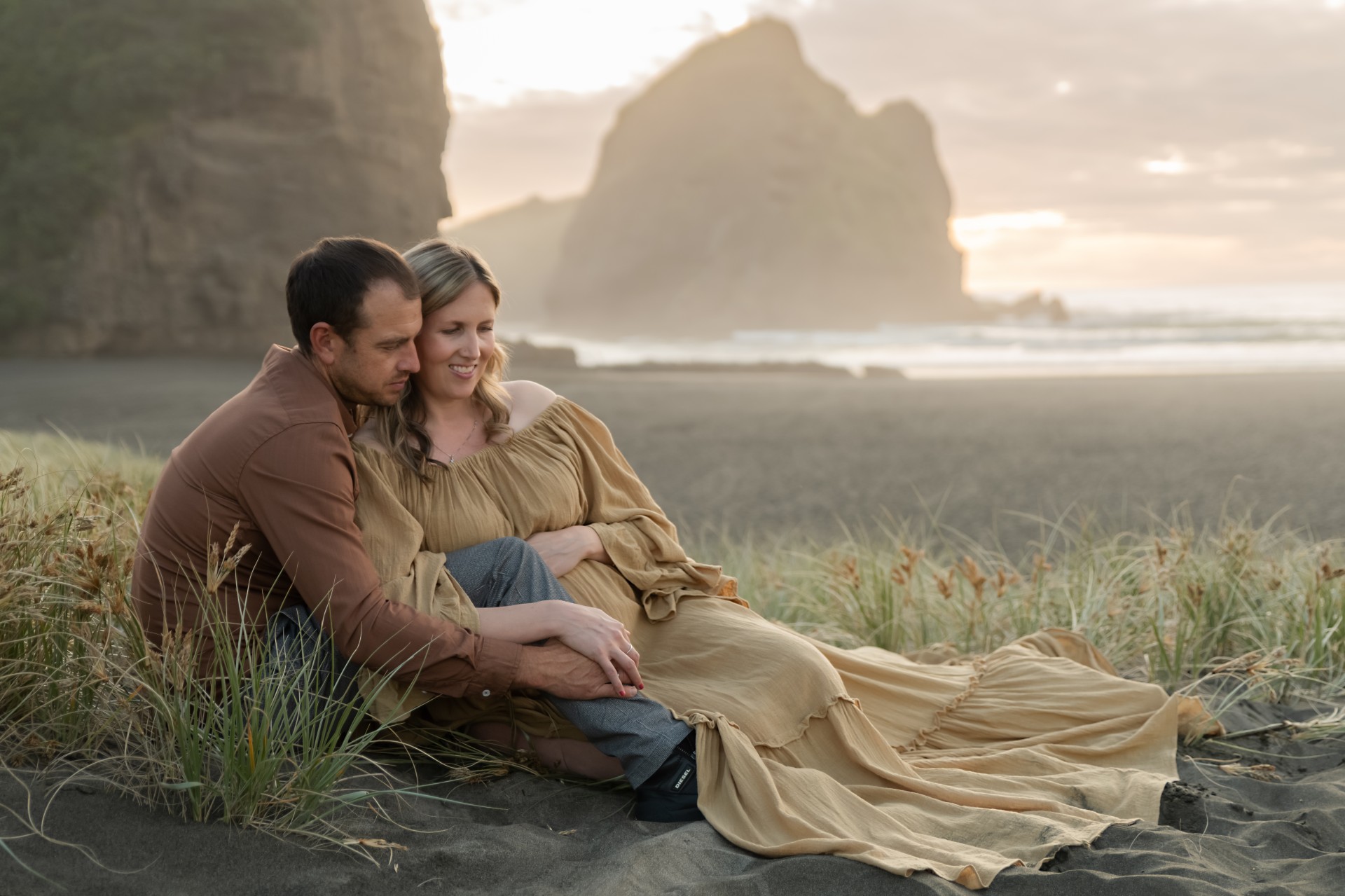 expecting couple is sitting in the long grass in piha beach during their maternity session