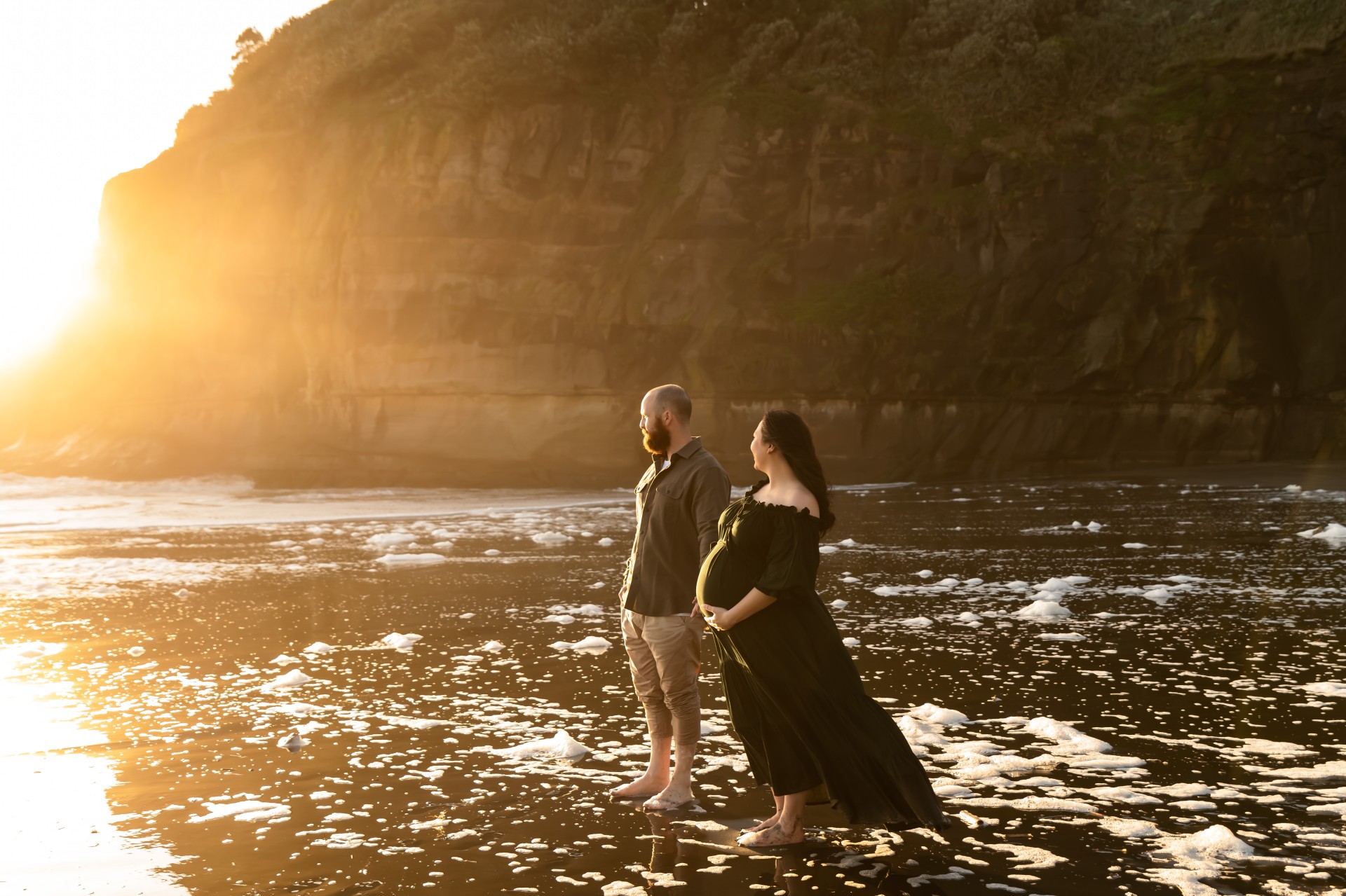 expecting couple is looking into the sunset on murriwai beach