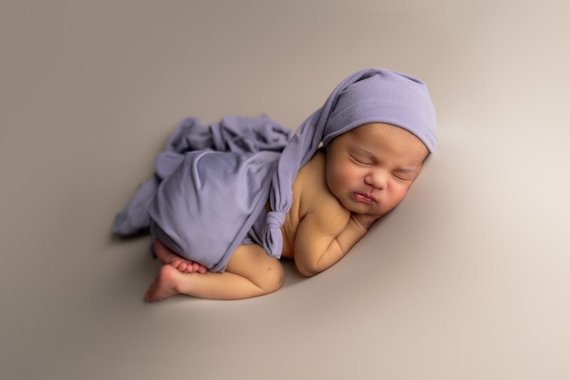 baby sleeping in the newborn bump up pose. She is wearing a purple sleepy hat and covered with purple jersey wrap