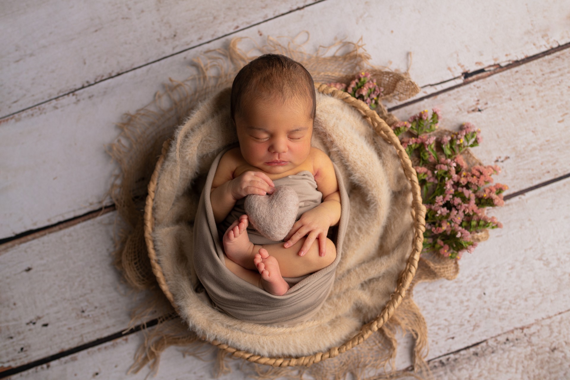 baby girl sleeping in the round basket, surrounded by flowers and holding a felted heart
